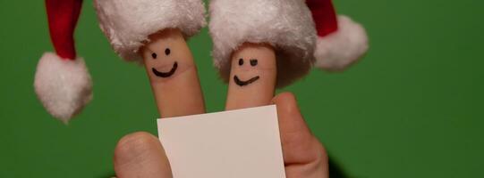 Fingers dressed in Santa-Claus red-white hats. Two happy finger smileys faces holding empty paper note for your text copy space against green background. Happy family celebrating concept for Christmas or New Years day. Finger couple Advertisement photo