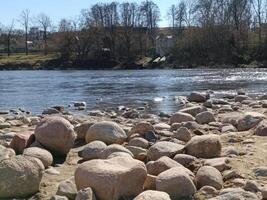 Smooth, rounded stones line the riverside, with a calm river and trees in the background, creating a serene outdoor scene. photo