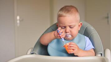 A one-year-old boy sits at a table and eats milk yogurt on his own. The child eats baby yogurt. The boy eats yogurt deliciously and smears it on his face. video