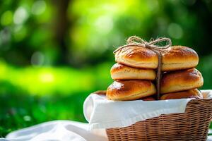 A stack of small brioche buns with golden-brown crust tied with twine, placed in a wicker basket outdoors on a white cloth, surrounded by a lush green bokeh background photo
