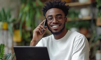 Young black man working talking mobile smartphone and on on laptop, smiling in home office with plants photo