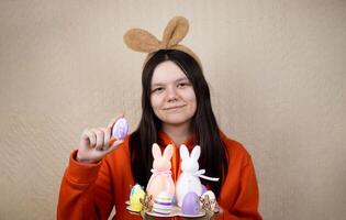 A pretty smiling girl in casual clothes with bunny brown ears, holding a stand with Easter eggs and bunnies, demonstrates a colorful egg on a simple background. Happy Easter Concept photo