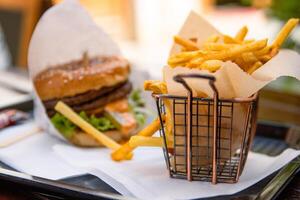 Closeup of a restaurant table with classic fast food meal. Burger with beef patty, fresh lettuce and a sesame seed bun wrapped in paper. Crispy golden French fries served on a tray. Selective focus 01 photo