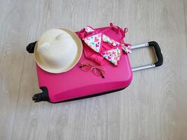 On the pink children's suitcase are a children's swimsuit, hat and sunglasses. A suitcase on wheels with a handle is lying on the floor. photo
