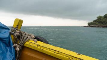 front of a brightly painted yellow boat with tied ropes. Against a backdrop of a calm gray sea and a distant. Rocky shoreline of island. Focus on the boat details and the transition to the open water. photo
