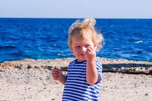 A young blonde child is standing on a beach, smiling and holding his hands up photo