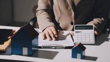 A woman in a business suit is writing on a piece of paper with a calculator and house model video