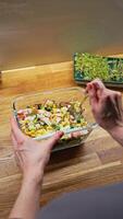 Vertical screen. female chef mixing surimi salad with corn, cucumber, and mayonnaise in transparent glass bowl on rustic wooden surface, highlighting culinary precision. POV shot. chef mixing salad video
