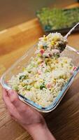 Vertical screen. hand lifting spoonful of surimi salad from a transparent glass bowl, surrounded by fresh ingredients including surimi, corn, cucumber, boiled eggs, and arugula on rustic wooden table video