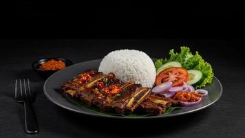 Delicious grilled beef ribs served with rice, sambal, and fresh vegetables on a rustic plate photo