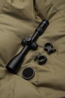 A black rifle scope with adjustment knobs, mounting rings, and a honeycomb filter placed on a textured khaki fabric surface. photo