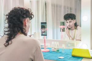 Young man filming himself applying makeup tutorial using smartphone on tripod in front of mirror photo