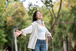 An asian woman is smiling while standing with open arms in the park, breathing in fresh air. photo