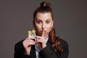Woman in business attire holding a yellow alarm clock, making a hush gesture, emphasizing time management, silence, and urgency. photo