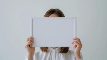 Woman in white blouse holds blank frame mockup in front of face. Minimalist neutral hidden female. Elegant simple template advertising image. Mock up branding product, marketing material photo