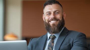 A well-groomed man with a beard and arm tattoos smiles while seated at a desk, dressed in a suit and tie, during a bright day at work photo