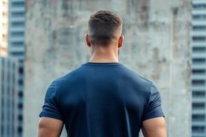 A muscular young man with a short haircut stands with his back to a weathered concrete wall, gazing into the distance under clear skies photo