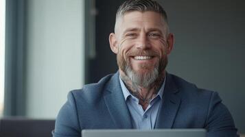 A white man with a beard and arm tattoos smiles happily while engaged with his laptop at a stylish office during daylight hours photo