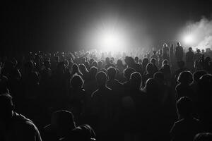 People fill the area at a nighttime event, illuminated by bright lights as they gather, creating a lively atmosphere in black and white photo