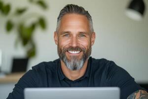 A white man with a beard and detailed arm tattoos sits at a desk, smiling while focused on his laptop in a well-lit office photo