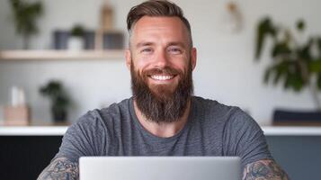 A white man with a beard and arm tattoos smiles while sitting at a desk, working on a laptop in a comfortable and inviting room photo