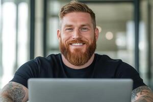 A white man with a beard and tattoos sits in a contemporary workspace, smiling while engaged with his laptop, creating a vibrant atmosphere photo