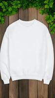 A plain white sweatshirt is draped over a wooden surface, with vibrant green leaves enhancing its display, creating a soft and inviting atmosphere photo