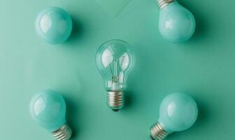 One clear light bulb emits a warm glow while four blue bulbs are arranged around it on teal photo