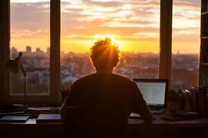 A person sitting at a desk with a laptop computer and a sunset in the background photo