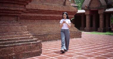Handheld shot, Solo female tourist enjoy listening to music from wireless headphones and playing smartphone while walking around the ancient site. Traveling and vacation concept video