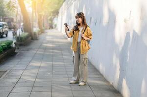 cheerful Asian woman in casual yellow jacket and cargo pants stands on sunny sidewalk, holding her phone and smiling while enjoying outdoors photo