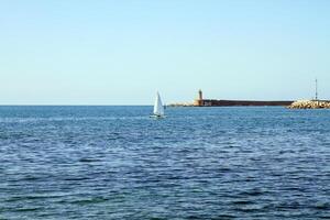 A sailboat in the open sea near a lighthouse and breakwater on a sunny day. photo