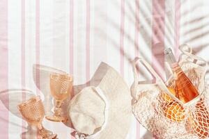 Summer Lifestyle flat lay with colored wine glass, bottle rose wine, sun hat, orange fruits in mesh bag on beach towel as background. Summer beach concept holiday and party, romance picnic photo