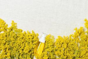 Cosmetic bottle container with yellow bedstraw herbs, wildflowers on natural textile table background, copy space. Blank label package for branding mock-up. Natural beauty product concept. photo