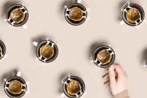 Pattern with black coffee cups, drip coffee bag with ground coffee for brewing in cup, paper bag drip coffee filter, female hand hold cup with drink, beige background, top view photo