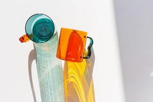 Orange and green Glass Mugs with bright sunlight Shadows, transparent cups casting dynamic, vibrant lights reflections and shadows on light white, creative aesthetic top view, minimal style, closeup photo