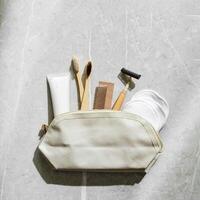 Beige fabric toiletry bag with eco-friendly personal care essentials on marble table. Bag with bamboo toothbrushes, wood comb, reusable razor, tube of natural cream or toothpaste, reusable pads photo