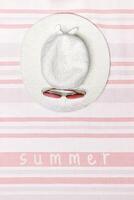 Minimal summer beach concept top view wide-brimmed sun hat with sunglasses on striped beach towel, pastel background. Summertime holiday or vacation card with text summer, pink white flat lay photo