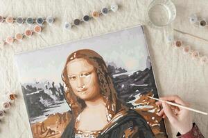 Painting by numbers with acrylic paints. Woman coloring Mona Lisa picture by numbers, hand with brush and paints of different colors. Creative hobby for relaxation, leisure activity at home photo