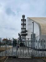 Industrial building with metal chimney and fenced perimeter, showcasing side view of rectangular structure with grey roofing and urban landscape in the background, under cloudy skies photo