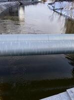Industrial pipes span across a river in winter, with snow-dusted banks and urban structures, serene waterway photo