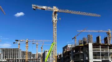 Tower cranes standing tall at a busy construction site against a clear blue sky. Multiple buildings in various stages of completion, modern architecture development photo