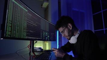 In a dark room illuminated by blue light, a young man focuses intently coding tasks. Two large monitors display streams of code and data as he navigates through programming challenges. video