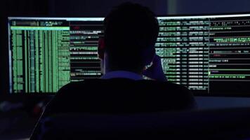 In a dark room illuminated by blue light, a young man focuses intently on coding tasks. Two large monitors display streams code and data as he navigates through programming challenges. video