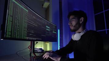 In a dark room illuminated by blue light, a young man focuses intently on coding tasks. Two large monitors display streams of code and data as he navigates through programming. video