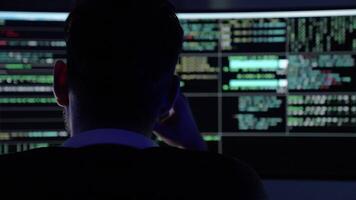 In a dark room illuminated by blue light, a young man focuses intently on coding tasks. Two large monitors display streams of code and data as he navigates through programming challenges. video