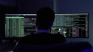 In a dark room illuminated by blue light, a young man focuses intently on coding tasks. Two large monitors display streams of code and data as navigates through programming challenges. video