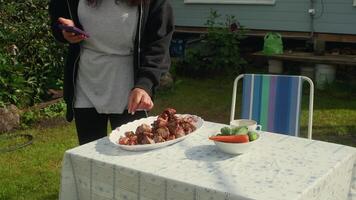 Woman is using her phone while preparing a barbecue of grilled meat in the garden, on a table with fresh vegetables such as cucumbers and carrots video
