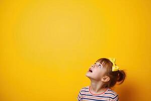 A young girl with a yellow bow in her hair looks up with a joyful expression against a bright yellow background. photo