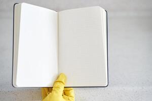 Yellow gloved hand holding open blank notebook on light granite surface with subtle dot grid pattern. photo